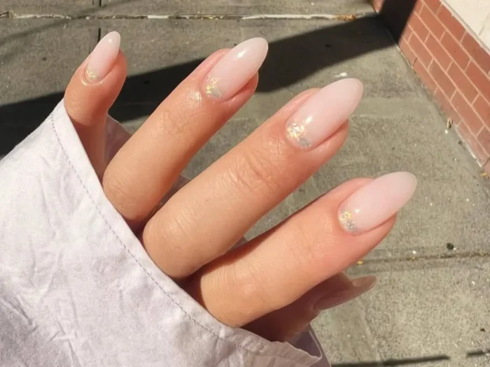 A close-up of a hand with a chic light pink manicure on medium-length almond nails, featuring a sheer milky pink base and subtle iridescent glitter flakes near the cuticles.