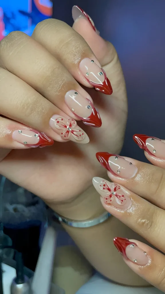 Stiletto nails with red V-French tips, sheer base, cuticle rhinestones, and red butterfly accent.