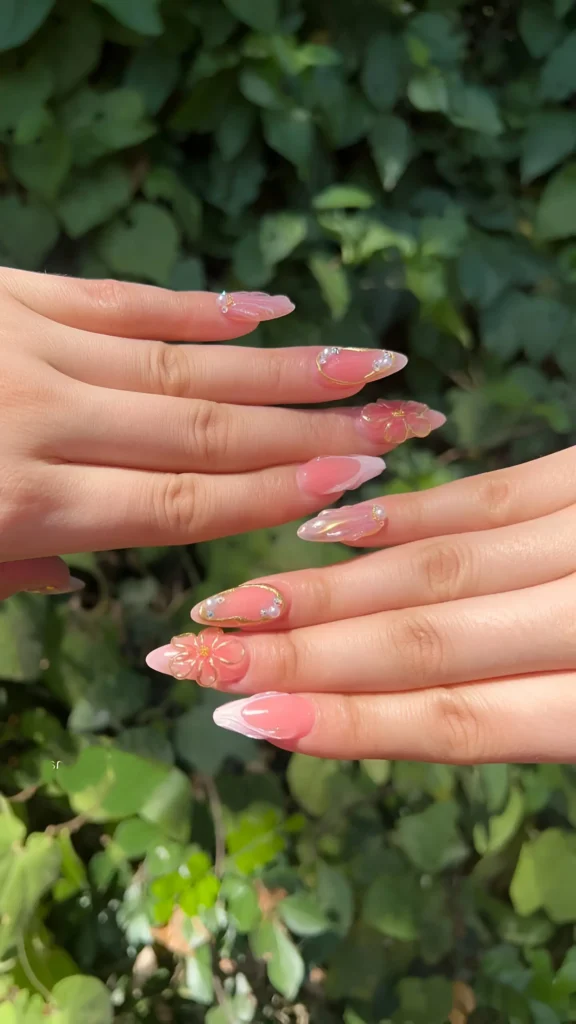 Almond-shaped summer nails with sheer pink base, 3D sculpted flowers, and gold accents.