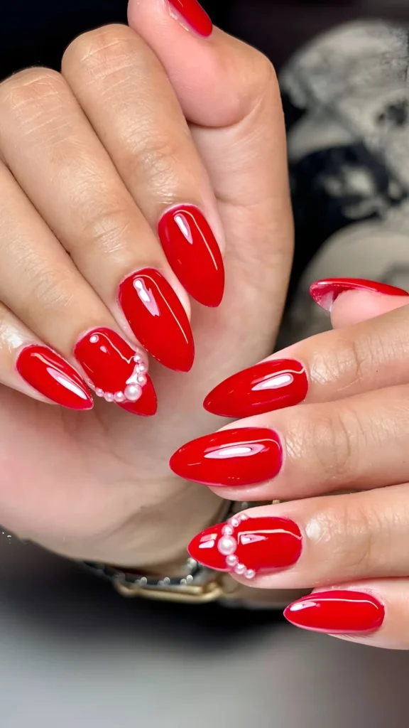Glossy bright red almond nails with pearl cluster accents near the cuticle.