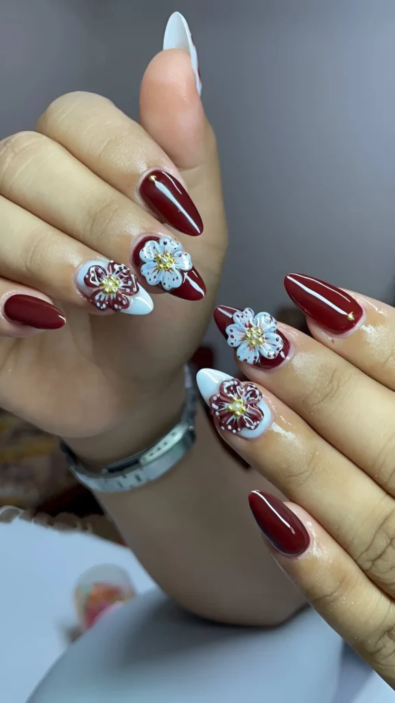 Almond nails alternating wine-red and white bases with contrasting 3D flower appliqués featuring gold centers.