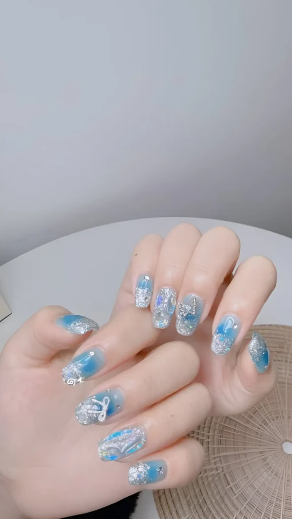 Short squoval nails with a sheer blue base, heavy silver glitter, and a white bow detail.