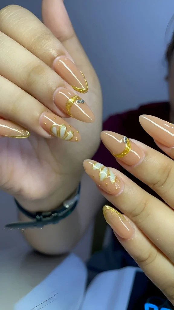 Stiletto nails with a creamy nude base, gold French tips, textured gold crescent moon details with crystals, and white floral accents.