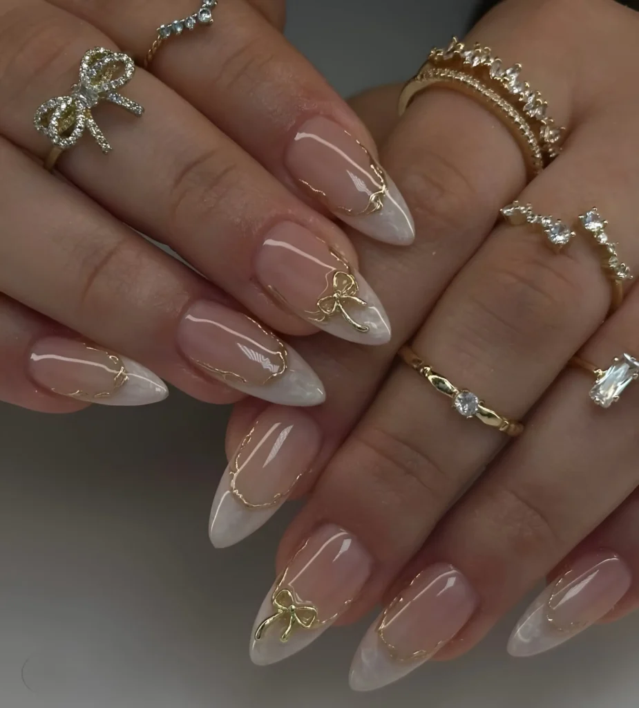 Delicate gold bow charms accenting a modern milky white French manicure on long almond nails.