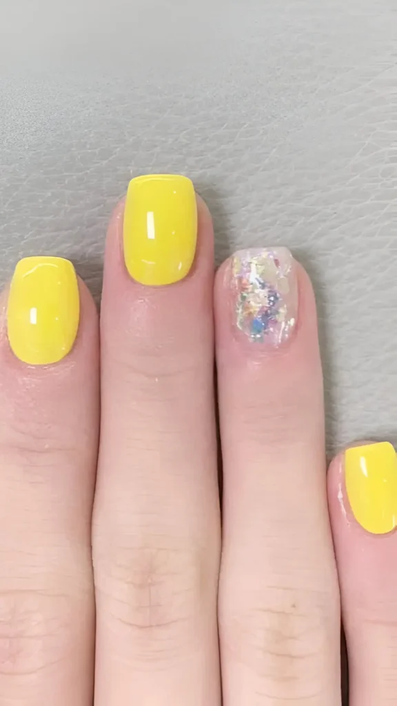 Short square nails with three nails painted solid bright yellow and one accent nail covered in chunky iridescent glitter flakes.