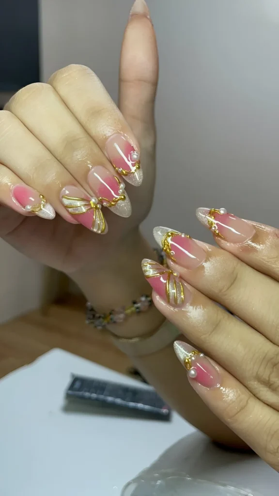 Pointed almond nails with a sheer pink blush base, white details, and 3D gold accents including bows with pearl centers and swirls.