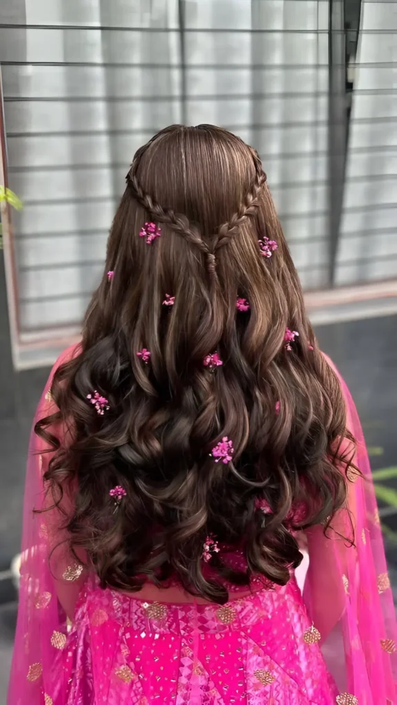 Back view of long wavy brown hair in a romantic half-up style with converging braids, adorned with small pink floral accessories.