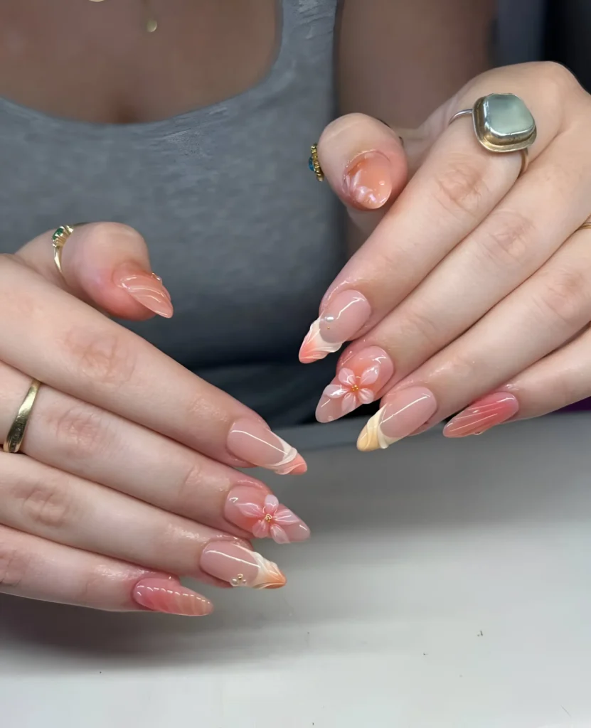 Almond nails with a mix of designs in warm peach and coral tones, including marbling, multi-color French tips, a 3D flower, and white swirls.