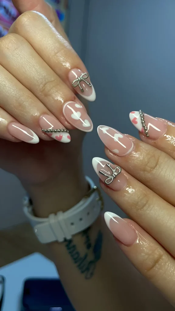 Almond nails with French tips, pink heart details on a sheer base, and 3D silver accents including bows and chains.