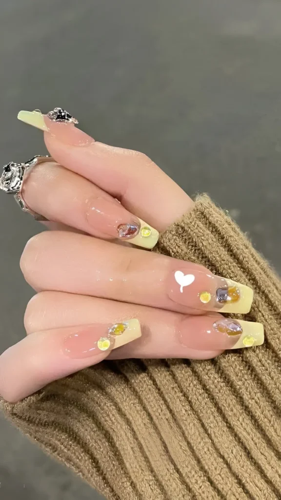 Long nails with sheer bases and pastel yellow tips, decorated with small white hearts, iridescent gems, and yellow glitter dots.