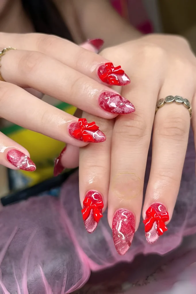 Pointed almond nails with sparkling red glitter polish and large 3D red bow embellishments.
