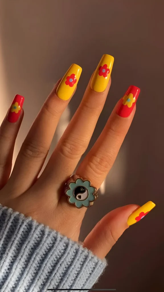 Long square nails with alternating bright yellow and red bases, each featuring a simple retro flower design in the contrasting color.