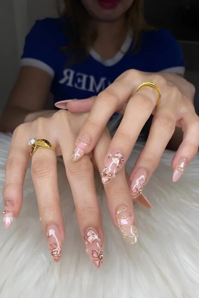Stiletto summer nails with sheer base, intricate gold outline patterns, pink accents, and small gems.