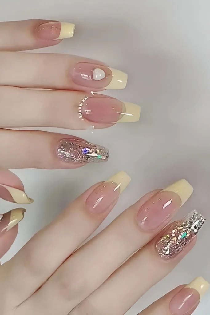 Long nails featuring pastel yellow French tips mixed with full-coverage silver/champagne glitter accent nails and details like a small white heart and rhinestones.
