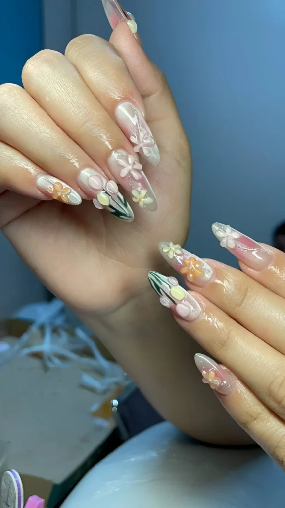 Long sheer stiletto nails adorned with scattered 3D pastel flowers (pink, yellow) and sharp green leaf-like graphic accents near the tips.
