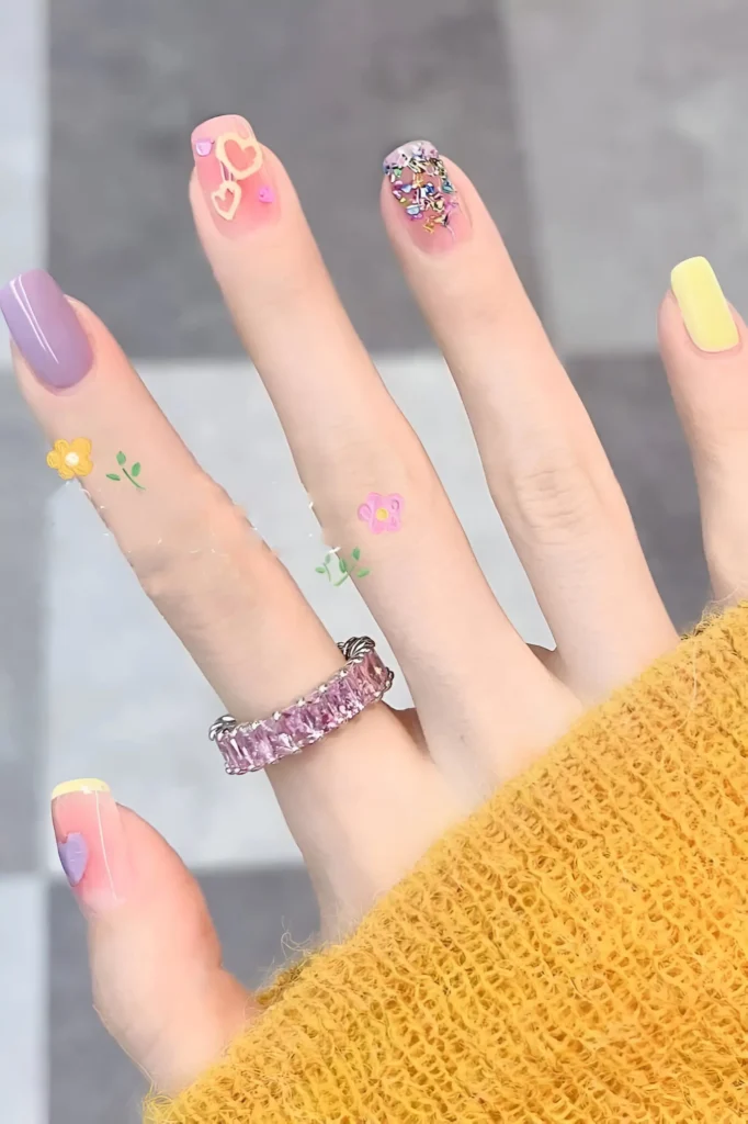 Short pastel manicure featuring solid yellow and lavender nails, a nail with yellow and pink heart outlines, and a nail covered in colourful microbeads or glitter.