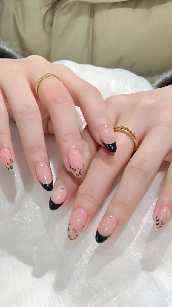 Hands displaying almond nails with a mix of black French tips and leopard print designs on a sheer base, accented with small rhinestones.
