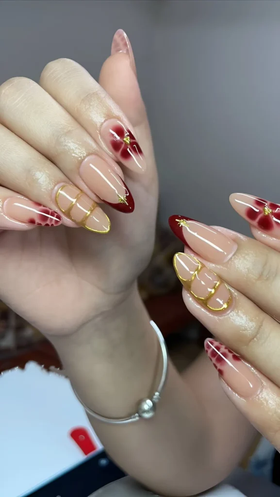 Mix-and-match stiletto nails with sheer nude base featuring red V-tips, watercolor red florals, and geometric gold line cages, all with gold star accents.