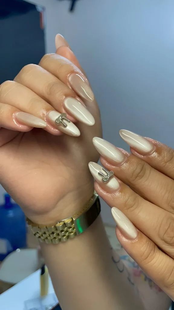 Almond-shaped nails painted with pearlescent white polish, featuring a single 3D silver bow charm on the accent nail.