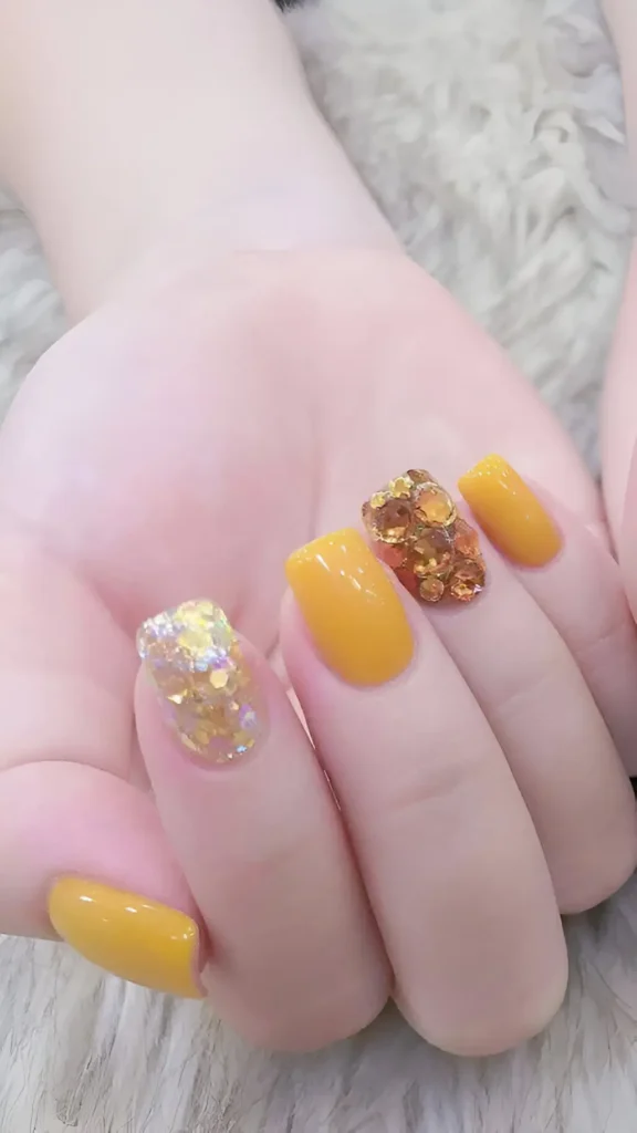 Medium square nails featuring solid marigold yellow polish mixed with accent nails: one with chunky iridescent gold glitter flakes, one covered in faceted golden yellow gems.