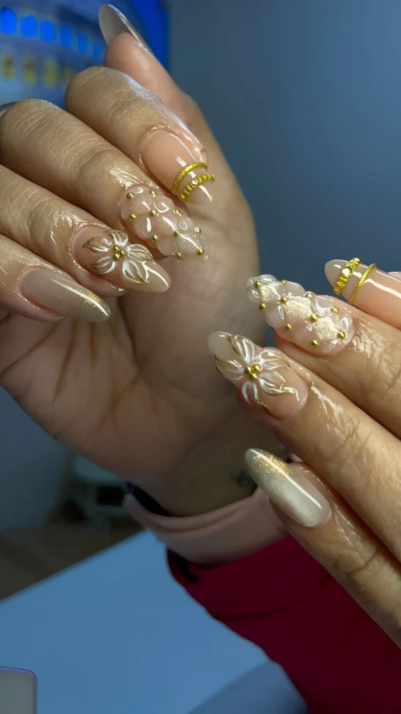 Almond nails with a sheer nude base featuring white florals, textured gold bead patterns, and gold line accents near the cuticle.