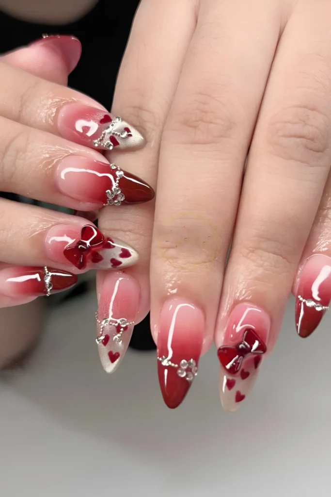 Pointed almond nails with red V-tips, scattered hearts, 3D red bows, and crystal chain embellishments.