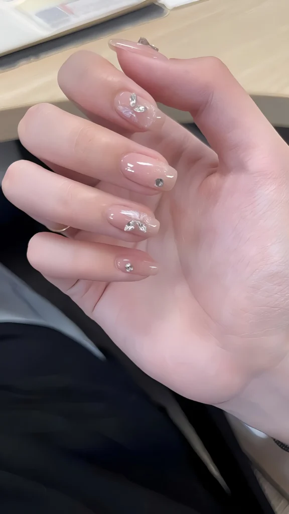 Hand displaying short-to-medium oval nails with a sheer nude glossy polish, decorated with small silver 3D butterfly charms and tiny rhinestone accents, a delicate example of cute short nail art.