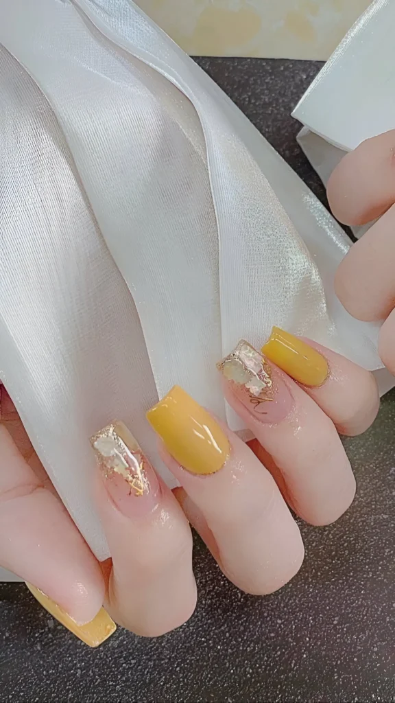 Medium square nails showing a mix of solid golden yellow polish and sheer nails decorated with crumpled gold foil accents.
