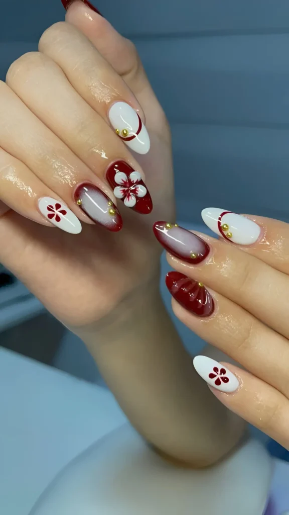 Almond nails alternating solid red and white bases, featuring graphic red and white floral motifs and abstract shapes accented with gold studs.