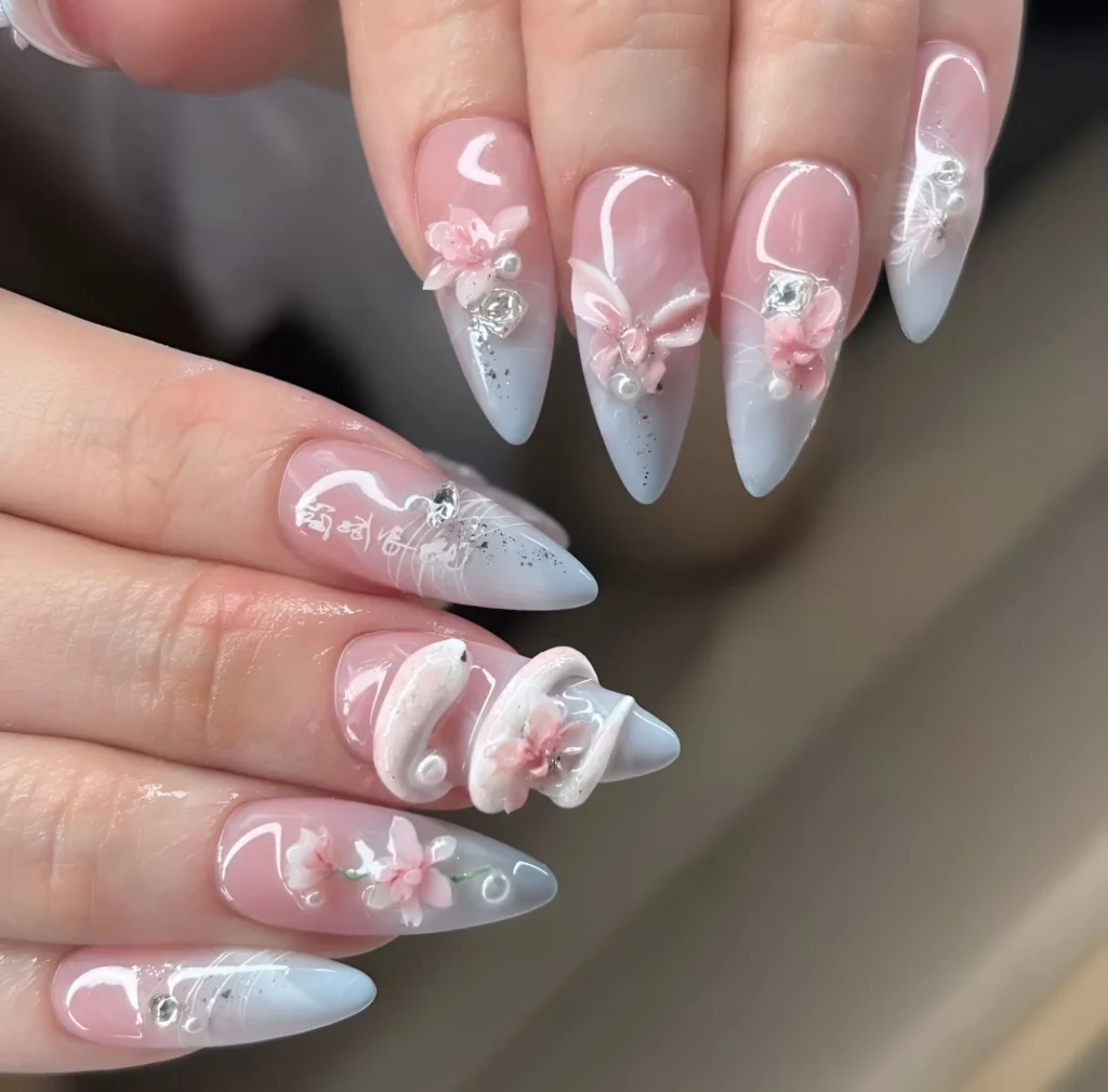 Long almond nails with sheer pink base, light blue hazy tips, 3D pink flowers, white sculpted swirls and bows, script, and gem accents.