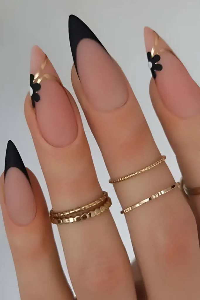 Long stiletto nails with sheer nude base featuring sharp black V-French tips and accent nails with black flowers and gold metallic details.