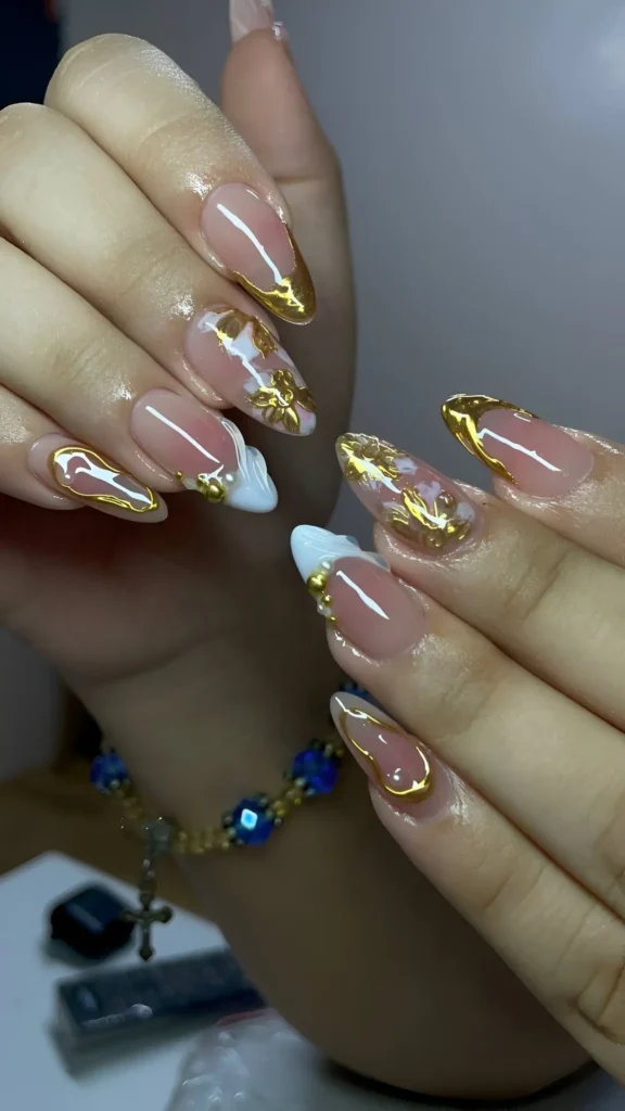 Almond nails with sheer nude base featuring abstract textured gold designs, dimensional milky white swirls, and gold bead accents.