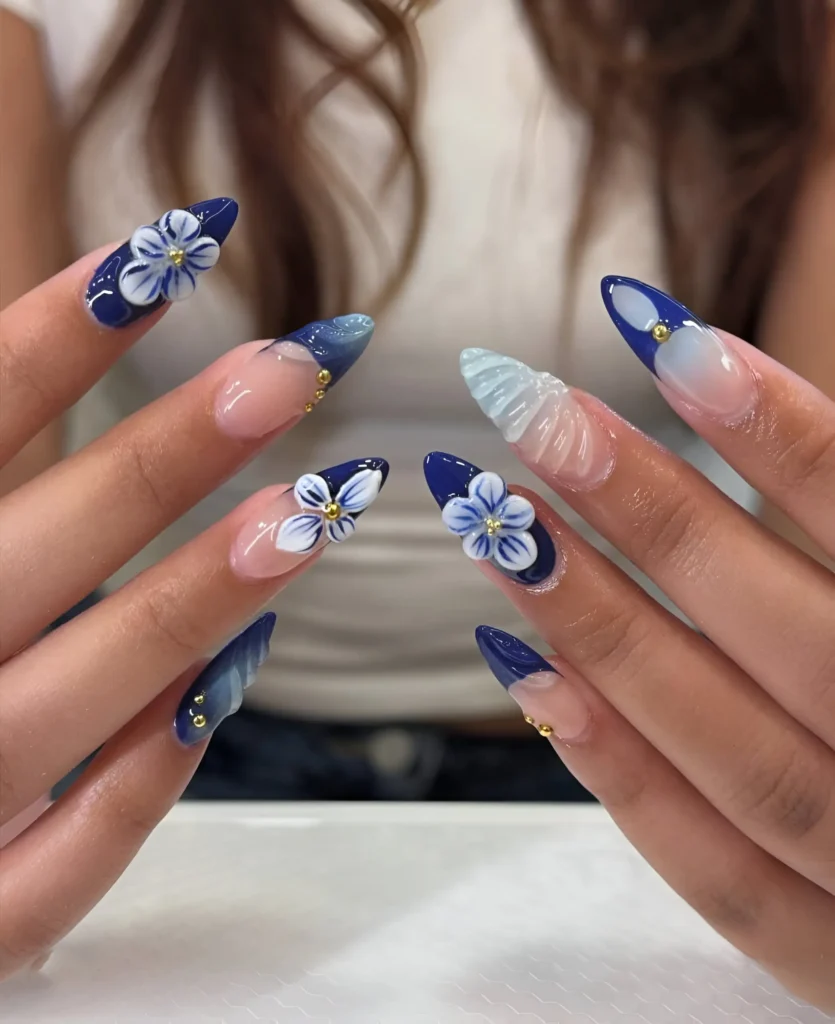 Almond nails mixing solid deep blue with white 3D flowers, blue French tips with gold studs, and a translucent textured accent nail.