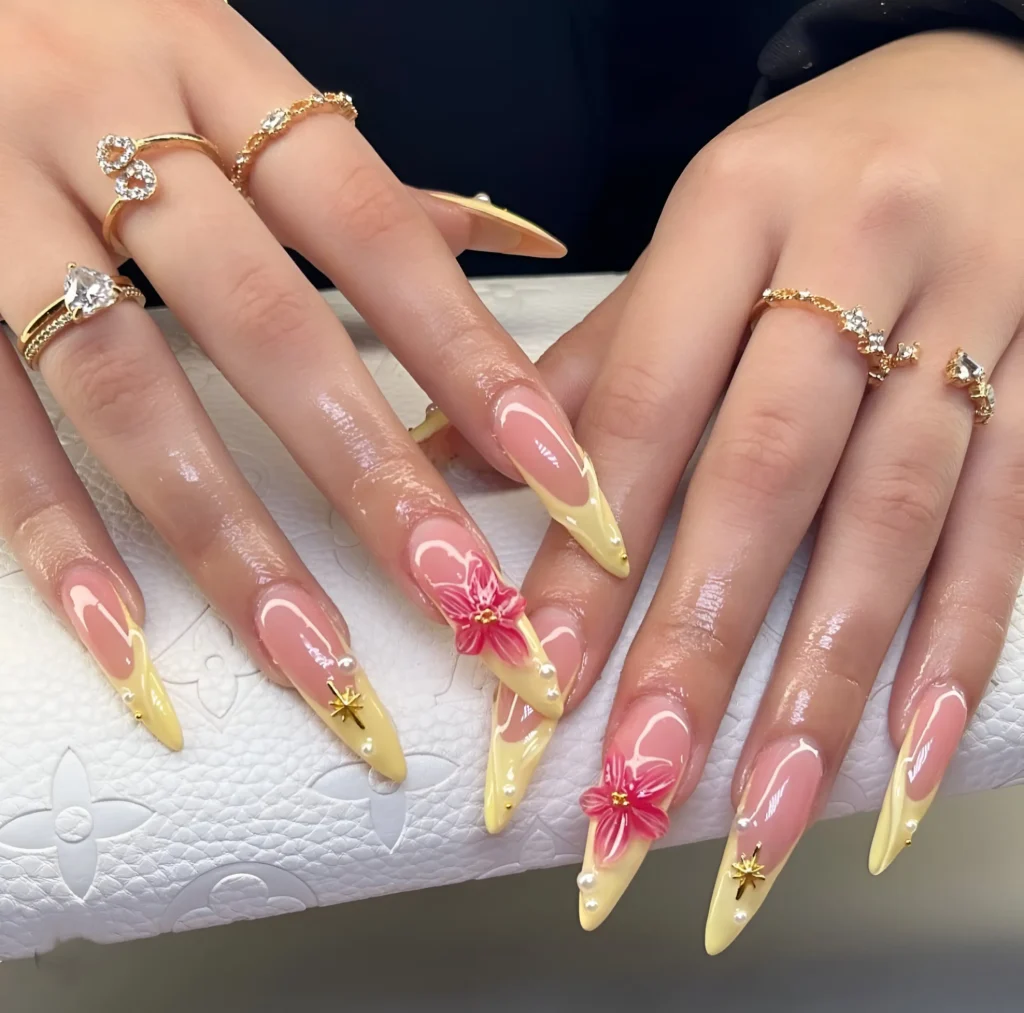 Long almond nails with pale yellow V-shaped French tips, 3D pink flowers, pearls, and gold star accents.