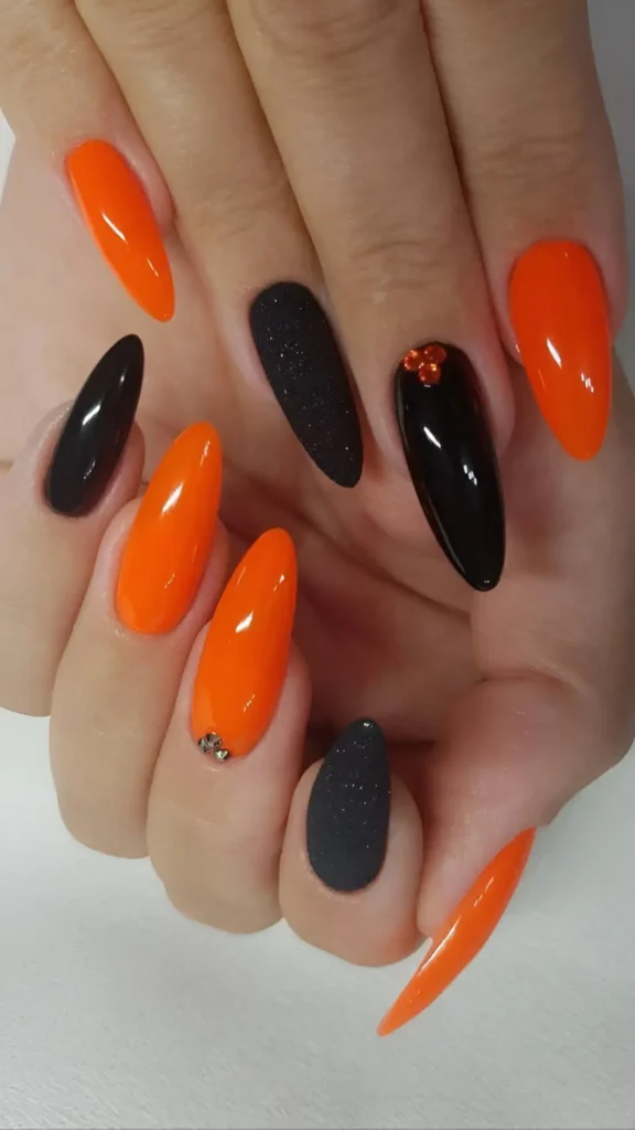 Almond-shaped nails with a mixed manicure featuring glossy bright orange, glossy black, textured black glitter, and rhinestone accents.