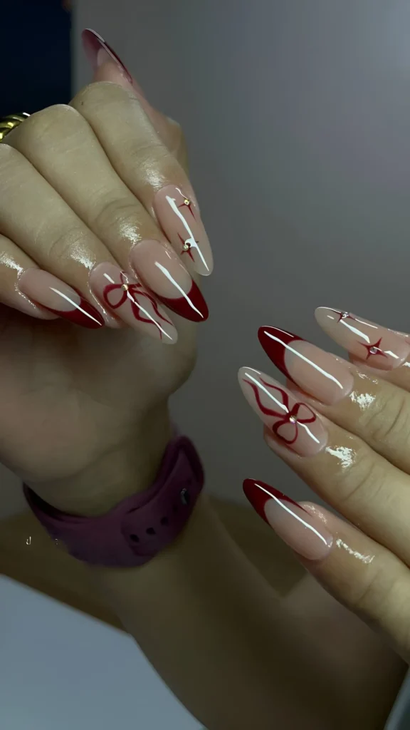 Almond nails with sheer nude base featuring deep red French tips, red line art bows, and tiny red starburst accents near the cuticle.