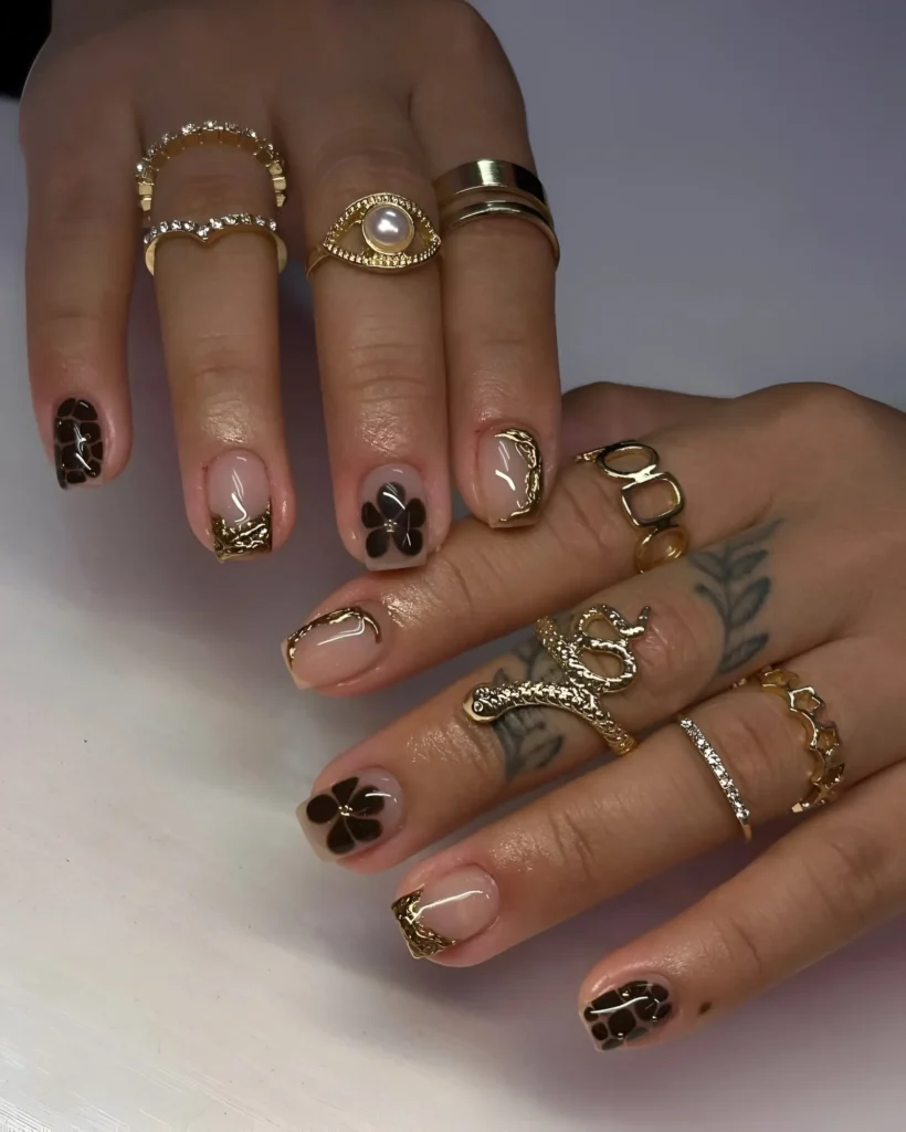 Short square nails with dark brown floral designs and textured gold accents over a sheer nude base.