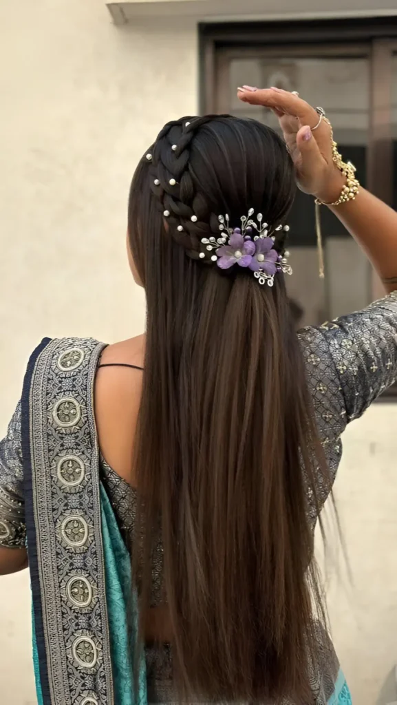 Back view of long straight dark hair in a half-up style with a pearl-accented headband braid and a large purple floral accessory.