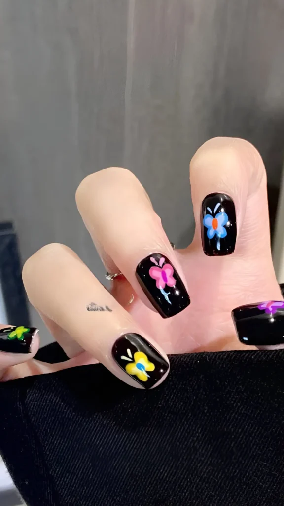 Hand with short square glossy black nails, each decorated with a simple, brightly colored flower design.