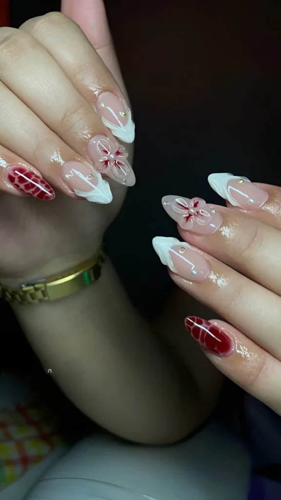 Almond nails with sheer nude base featuring white angular shapes, red floral motifs, sparkle clusters, and a deep red textured accent nail.