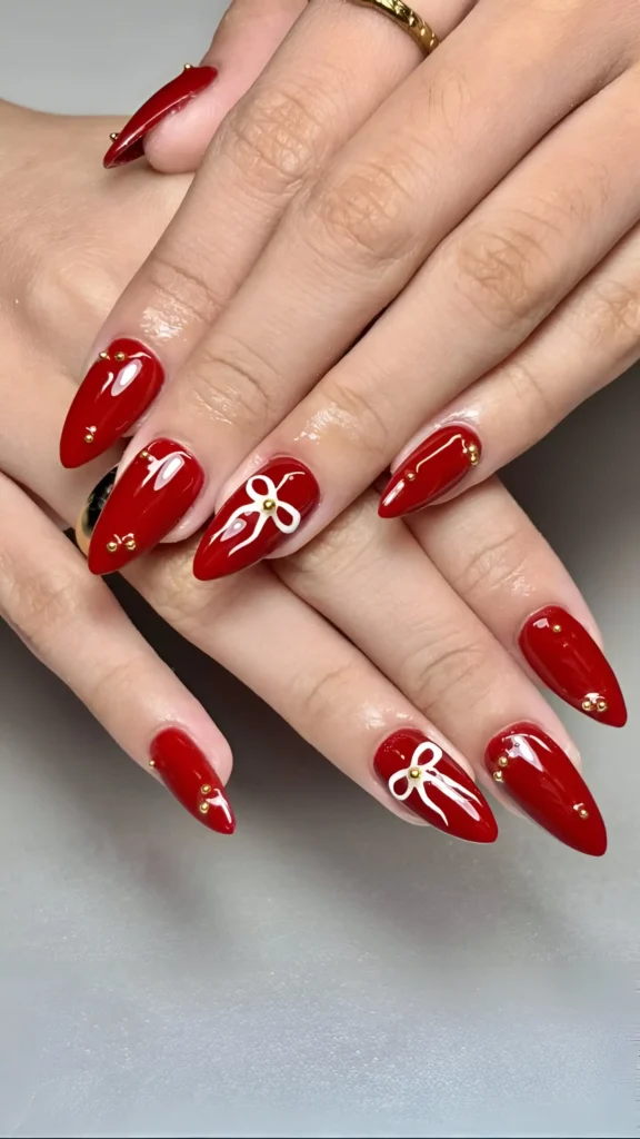 Pointed almond nails in deep red polish with white bow outlines centered with gold studs and scattered gold stud accents.