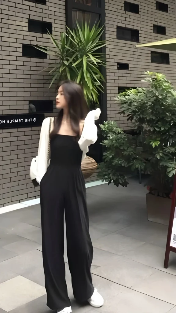Woman wearing a black square-neck wide-leg jumpsuit layered over a white blouse with voluminous long sleeves, with white sneakers and bag outdoors