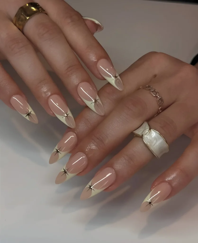 Long almond nails with sheer nude base, sharp white V-shaped French tips, and a metallic starburst charm at the point.