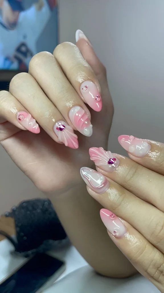 Almond nails with pink and white ombré base featuring dimensional sculpted waves/shells in pink and white, accented with pink heart gem clusters.