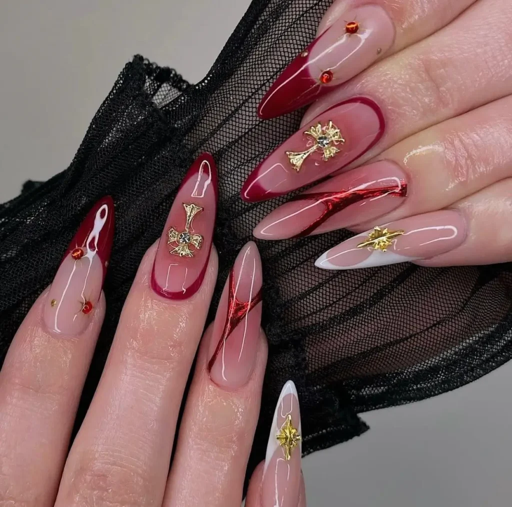 Long almond/stiletto nails with sheer base, deep red tips/outlines, white accents, metallic red lines, and large ornate gold charms (crosses, starbursts).