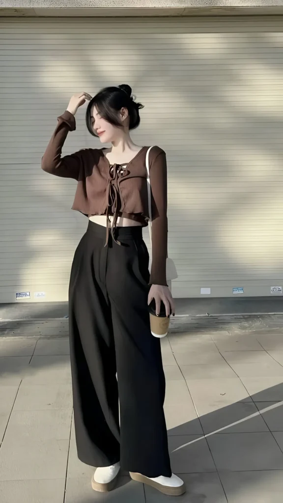 Woman wearing a brown cropped tie-front long-sleeved top, black wide-leg trousers, and platform sneakers, carrying a white bag and coffee cup outdoors