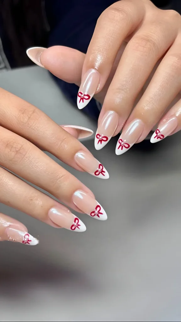 Long pointed nails with white French tips on a pearlescent base, detailed with small red bow outlines.