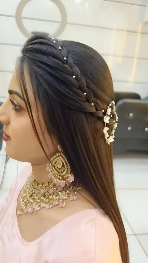 Side view of long straight dark hair with a pearl-lined headband braid along the hairline, finished with a large white floral accessory.
