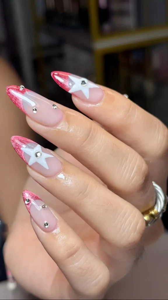 Long stiletto nails with pink glitter V-tips, white star outlines with gem centers, and scattered rhinestone accents.