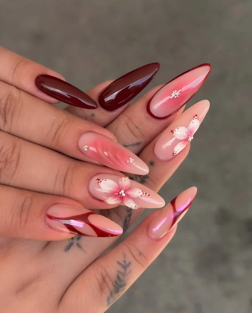 Stiletto nails mixing pink aura blush effects, painted white florals, burgundy accents, and red chrome swirls.